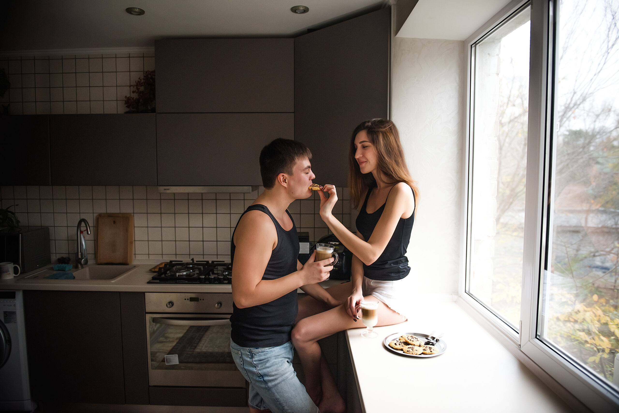 A young millennial couple is sitting on the windowsill near the window, drinking latte coffee with cookies in the kitchen at home in the apartment. Lovers hug, kiss, have fun. Woman treats sweet man
